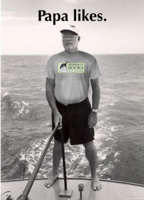 Papa Hemingway is standing on the deck of a sail boat out at sea. He appears to be steer the sail. And he's wearing a tee shirt with the Monroe Street Books logo on it.